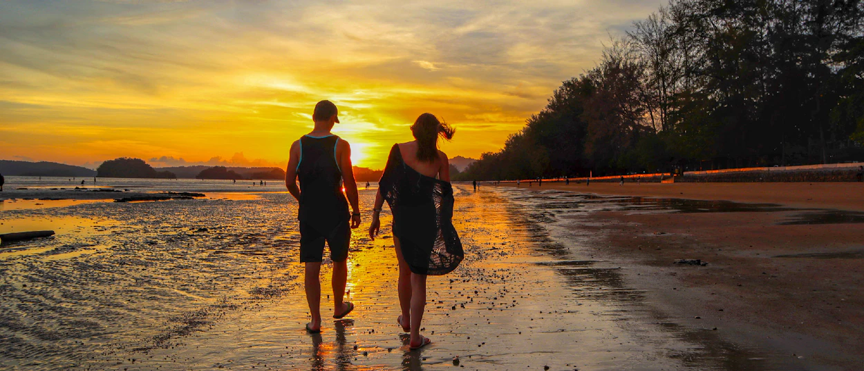 A couple walks along a wet beach at sunset in Krabi, Thailand, under a vibrant orange sky.