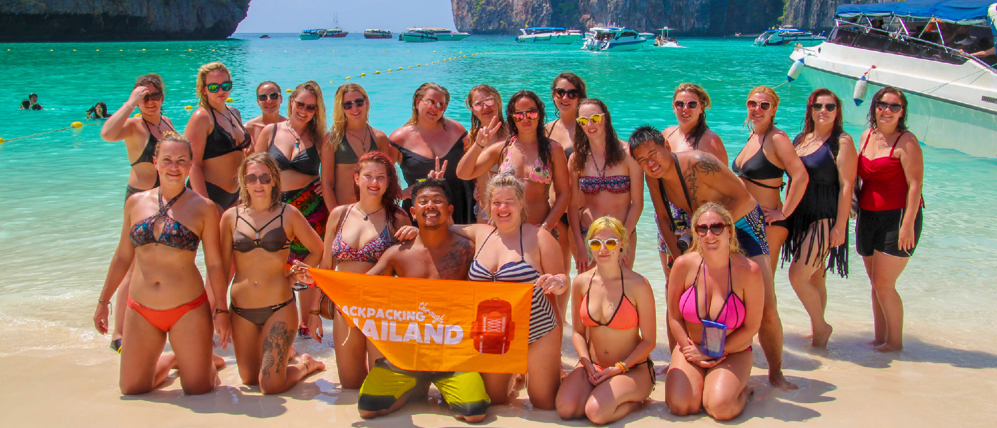 Backpacking group posing on a sandy beach with turquoise water, holding a tour bannerScenic turquoise water passage between lush limestone cliffs