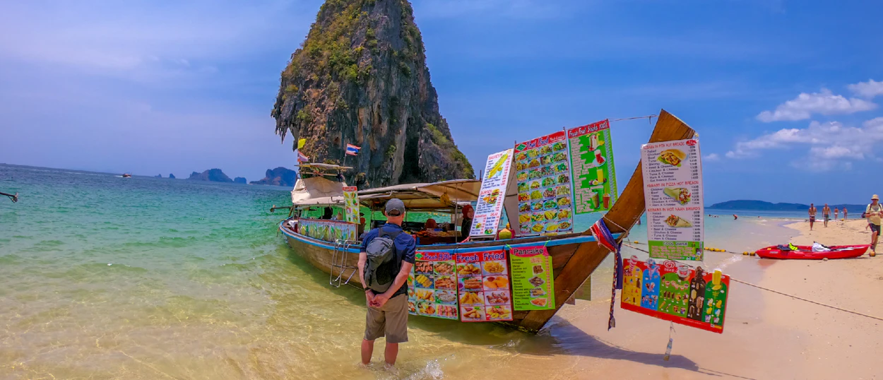 A man stands in clear water looking at a colorful longtail boat selling food on Railay Beach, Thailand.