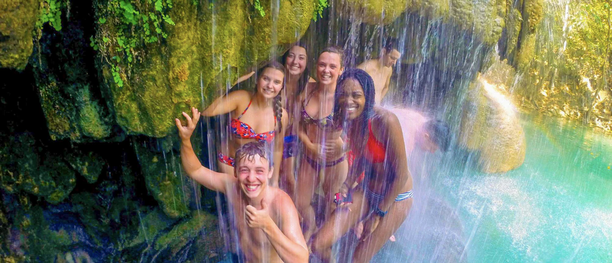Seven joyful travelers cool off under the emerald waters of a waterfall in Kanchanaburi, Thailand.