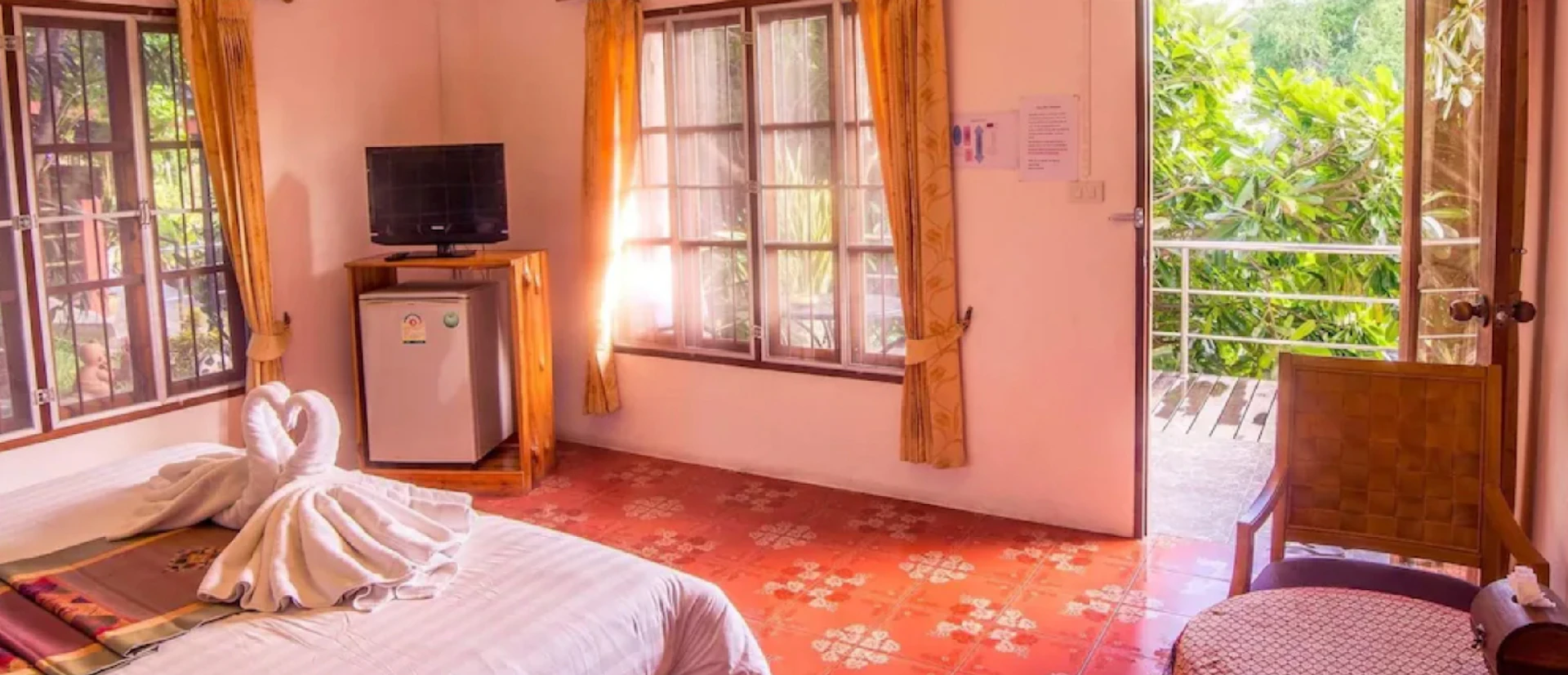 Bright hotel room with a bed, towel art, TV, mini-fridge, and a balcony overlooking trees.