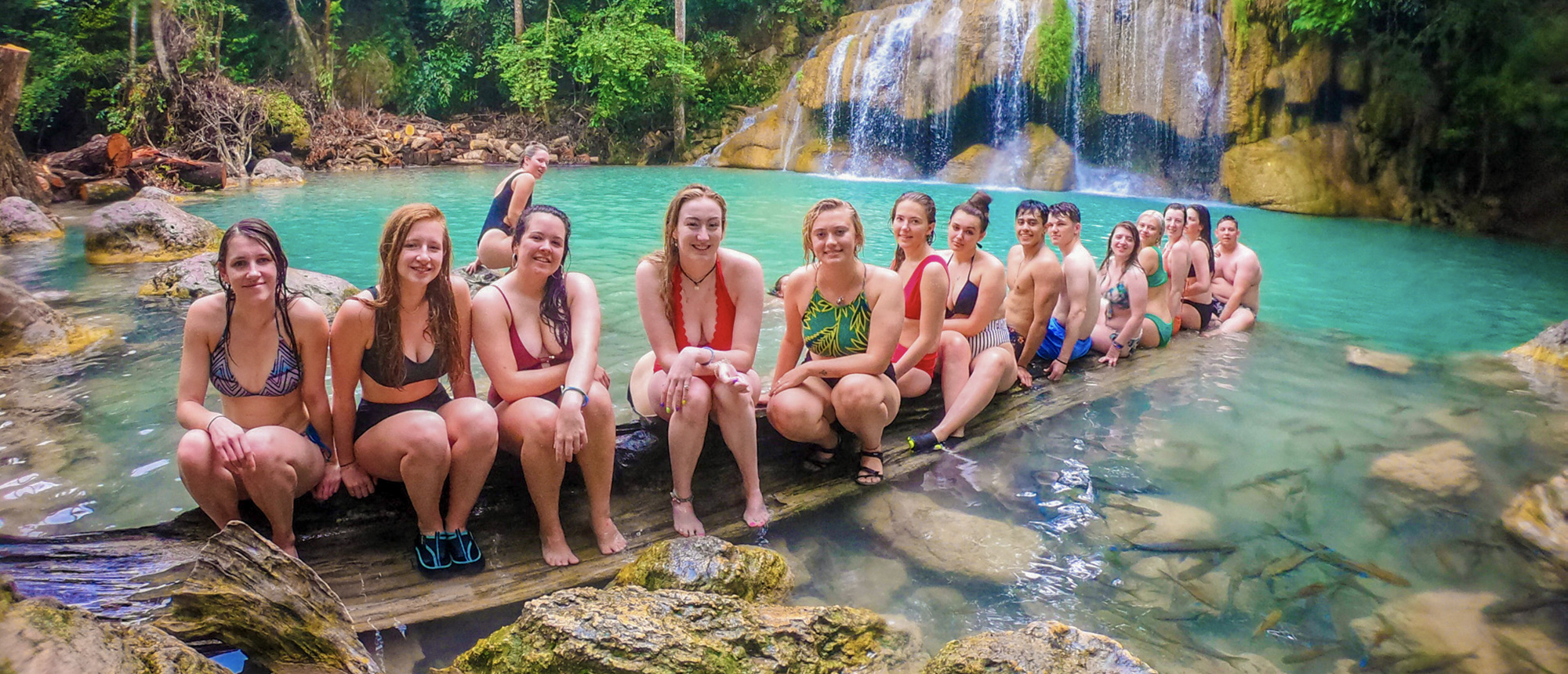 Backpacking group relaxing on a wooden deck beside an emerald pool at Erawan Waterfalls