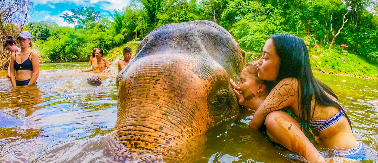 Two women lovingly kiss an elephant during a joyful river bath at a sanctuary in Kanchanaburi.