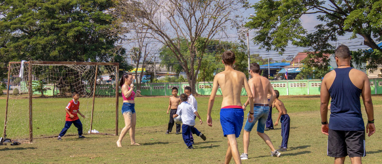 Backpackers and local Thai children happily play football together on a sunny school field in Amaphawa.