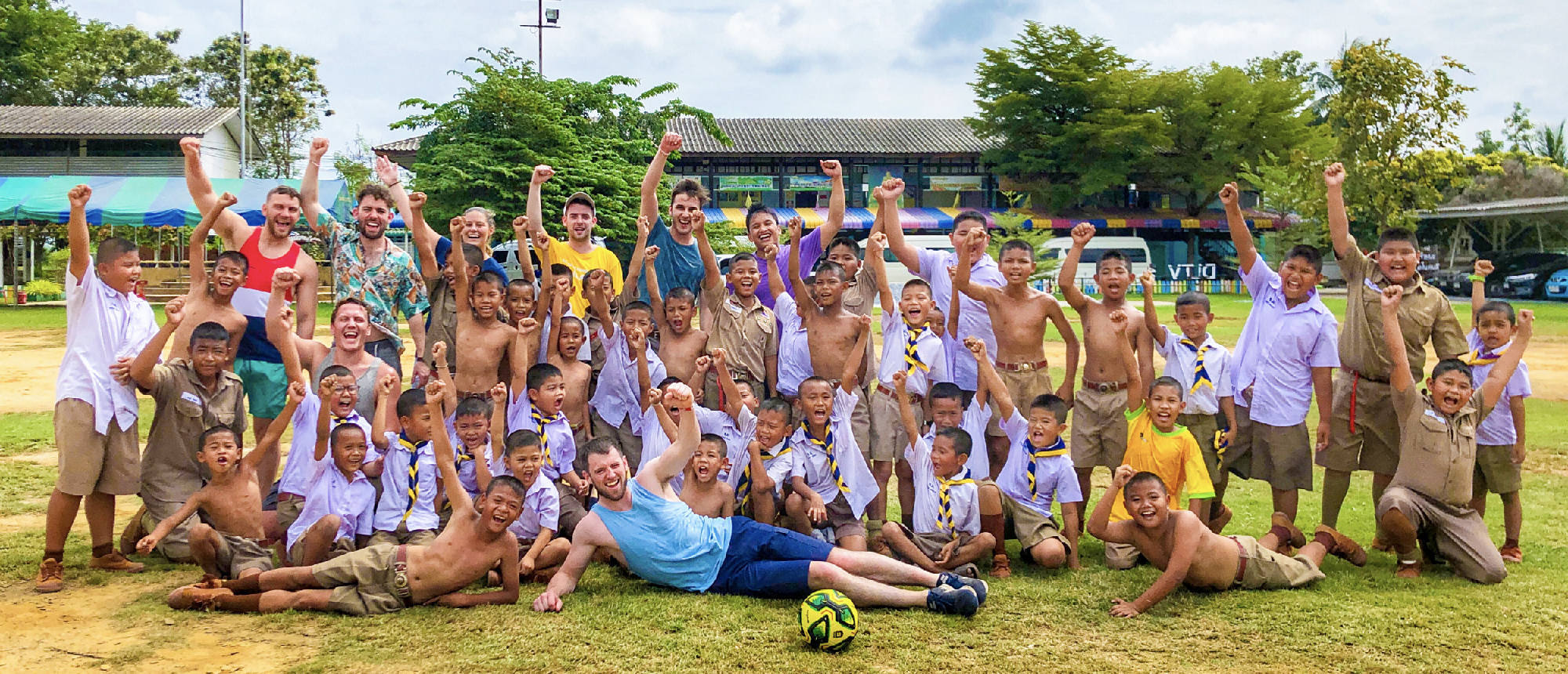 Backpacking group posing with local schoolchildren on a grassy field outside a Thai school