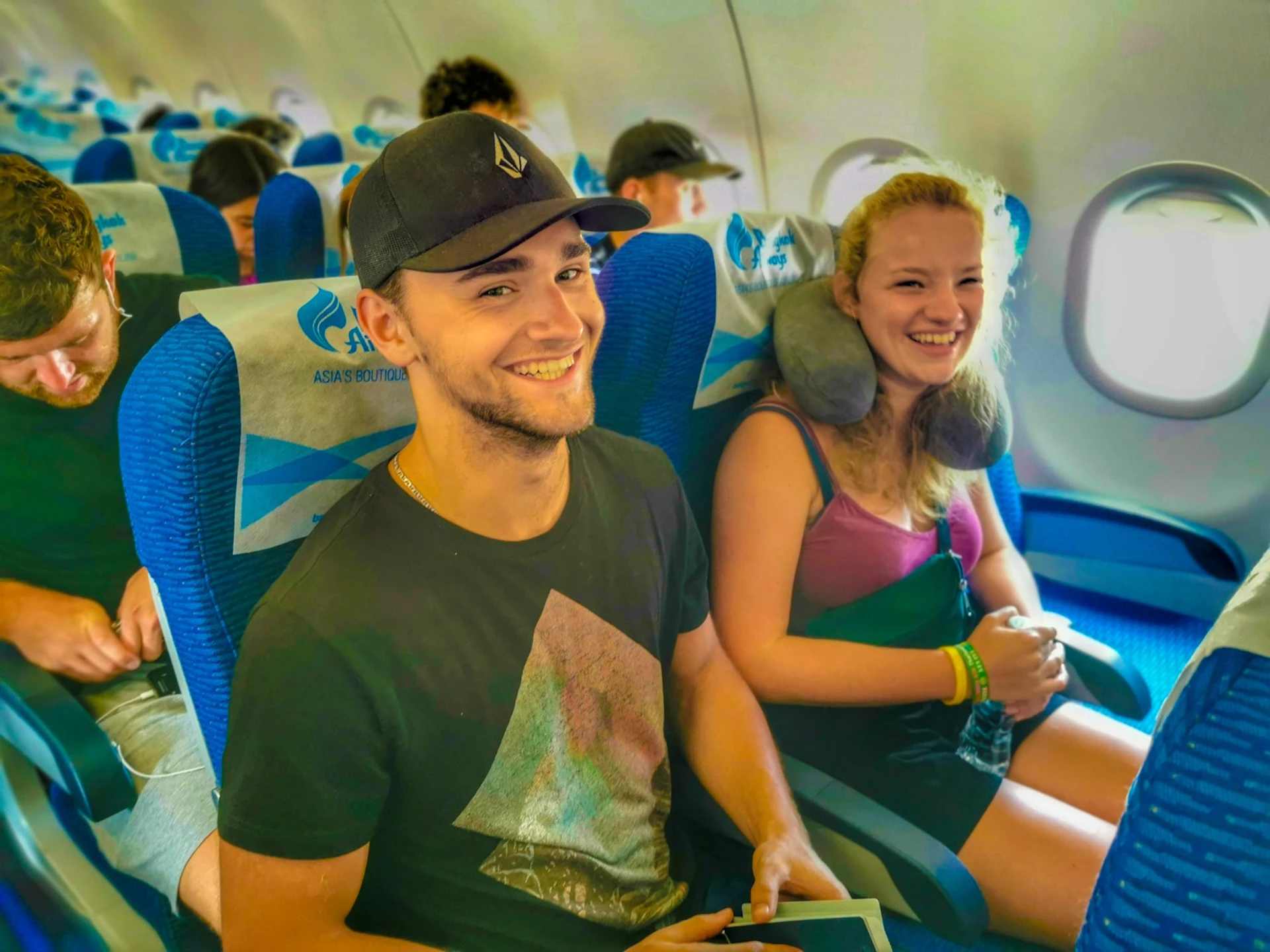 Two happy young backpackers smiling in blue airplane seats during a long-haul flight.