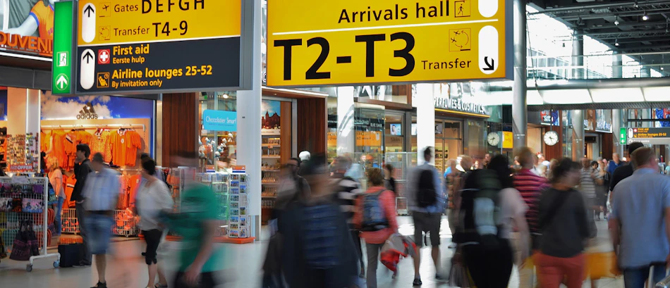 Travelers with backpacks move through a busy airport terminal with yellow directional signs.
