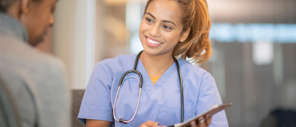 A smiling healthcare professional in blue scrubs with a stethoscope consults a patient, holding a clipboard.