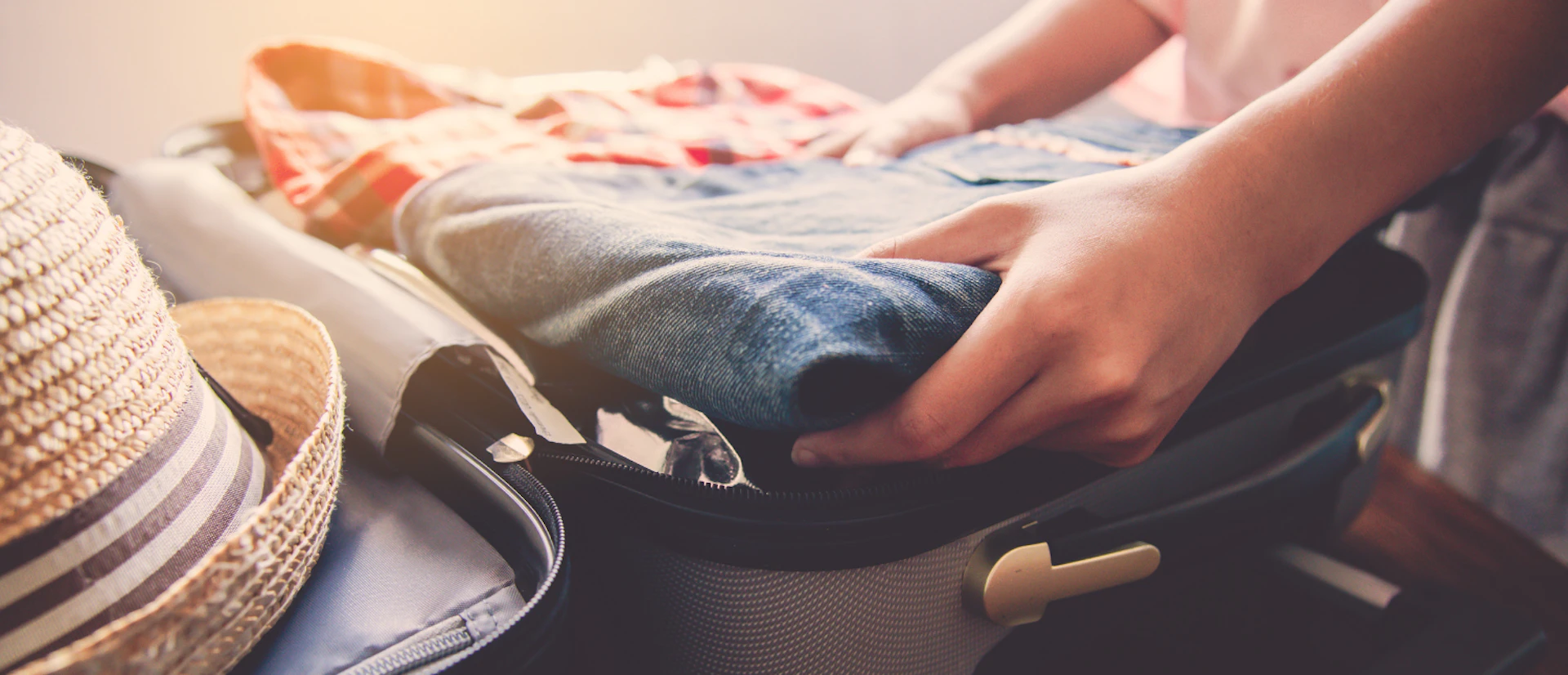 Hands carefully packing a suitcase with folded clothes and a straw hat, preparing for a tour.