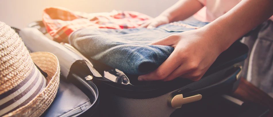 Hands carefully packing a suitcase with folded clothes and a straw hat, preparing for a tour.