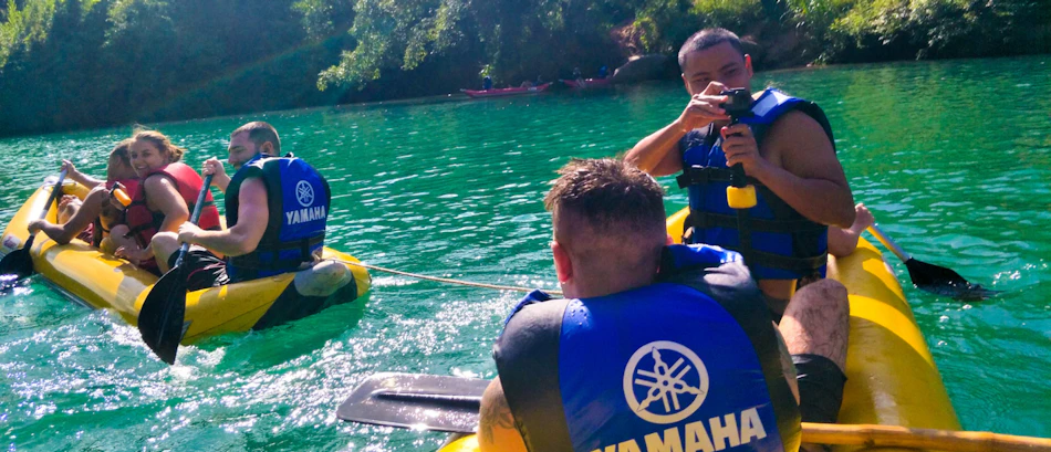 Friends in yellow kayaks with blue life vests on a clear turquoise river, one taking photos.