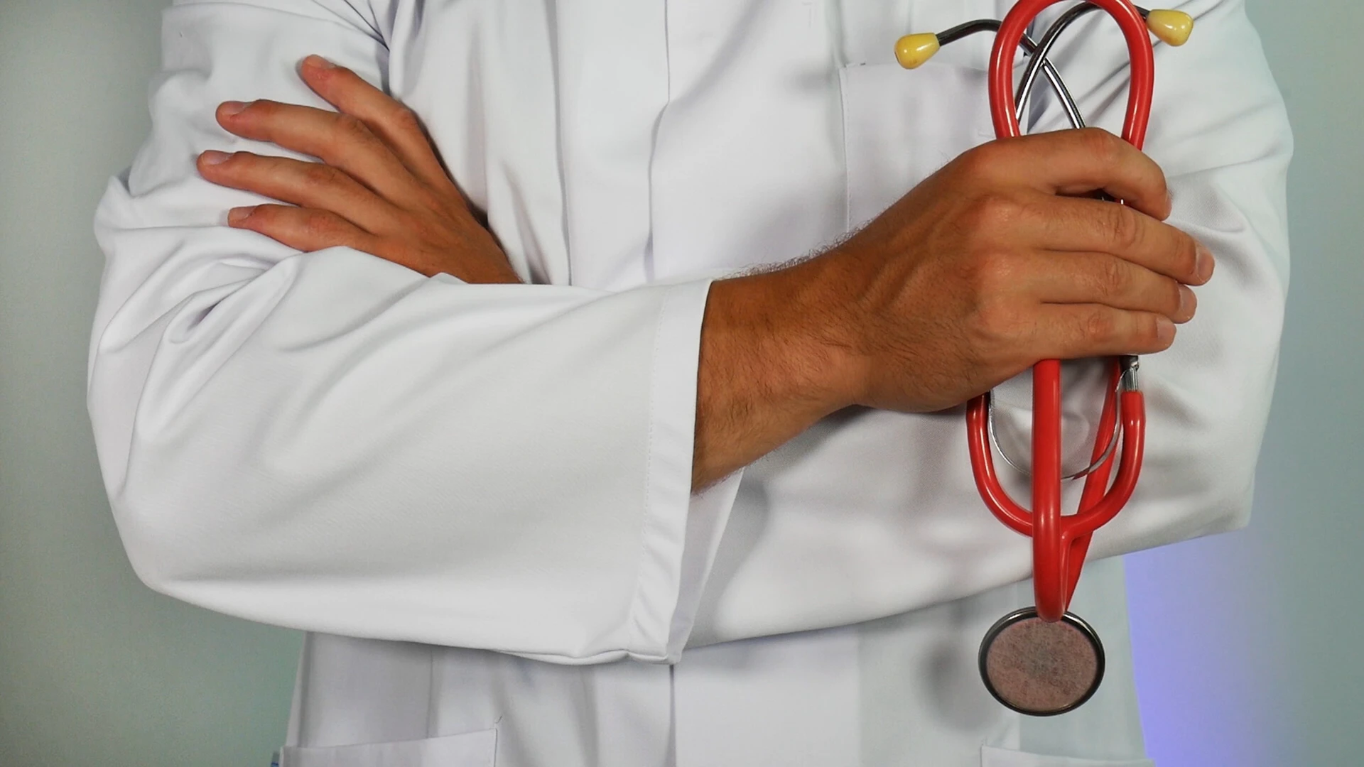 A medical professional in a white lab coat with crossed arms, holding a red stethoscope.