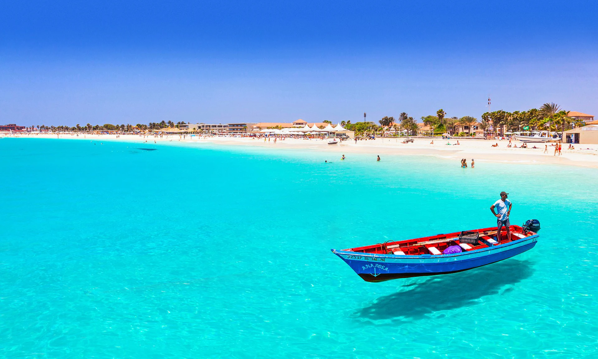 Man in a small boat on vibrant turquoise water near a white sand beach in Cape Verde.