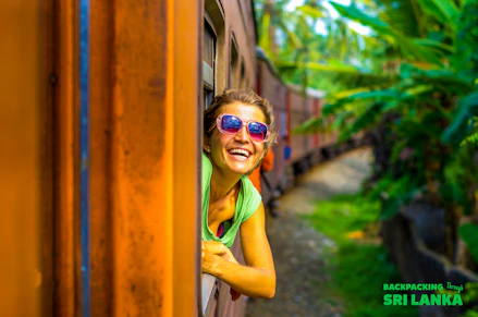 Smiling woman in pink sunglasses enjoying a scenic train ride through the tropical landscape of Sri Lanka.