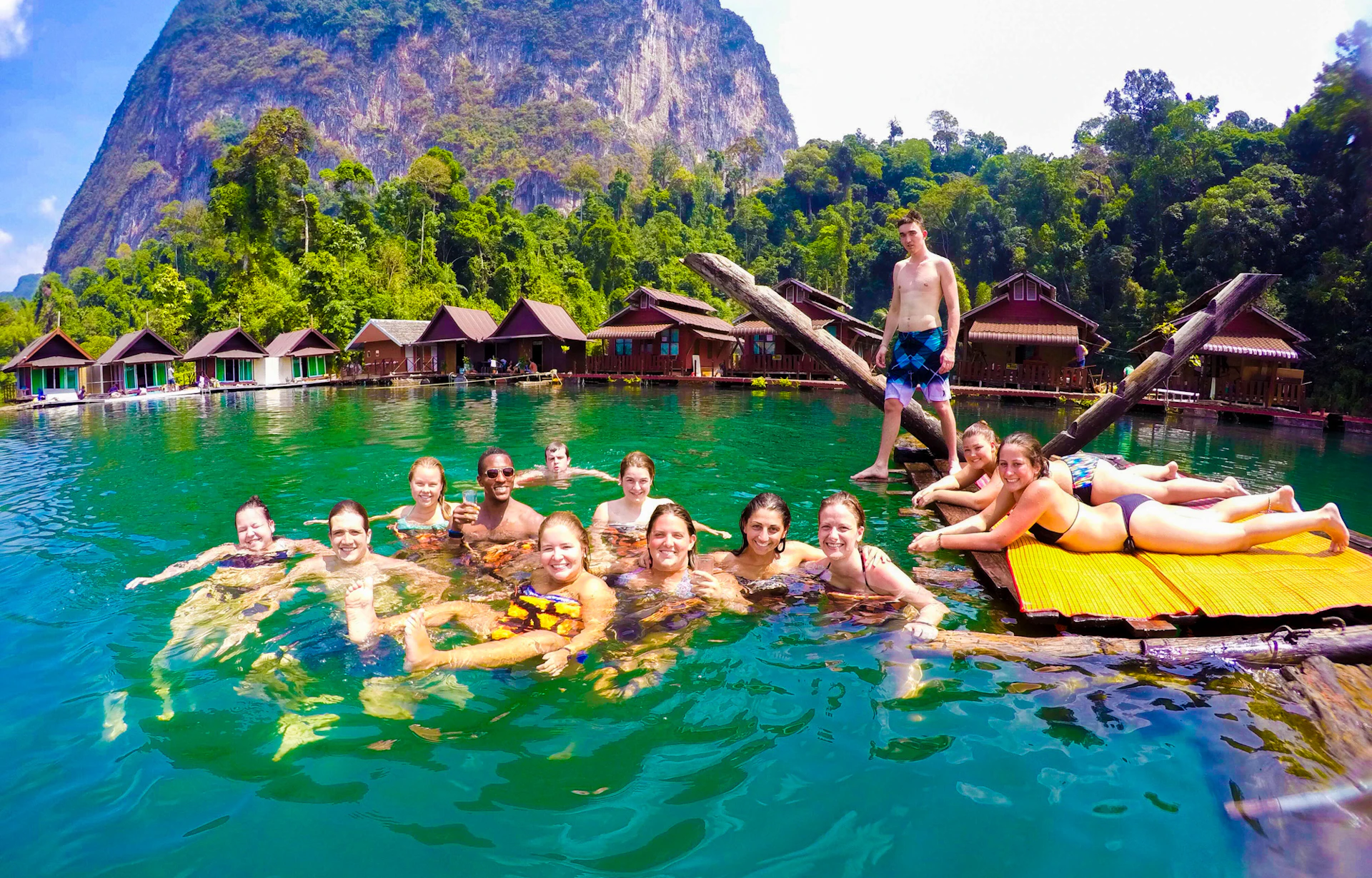 Happy backpackers swim and relax in the emerald waters of a Thai lake with floating bungalows.
