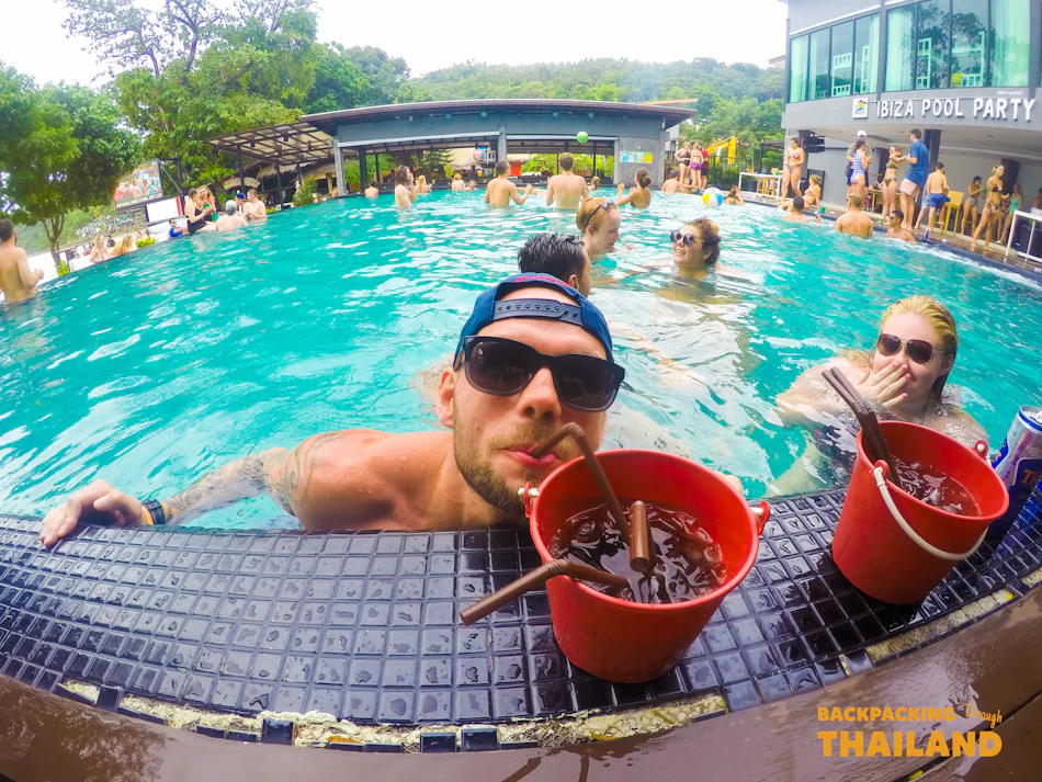 A man and woman enjoy bucket drinks at a crowded, lively pool party.