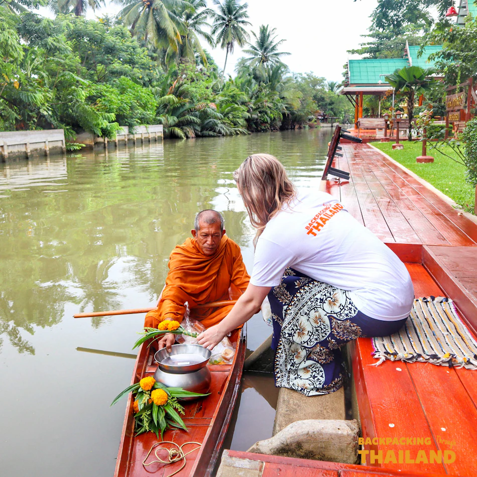 A woman offers alms to a Buddhist monk in a boat on a tranquil canal in Thailand.