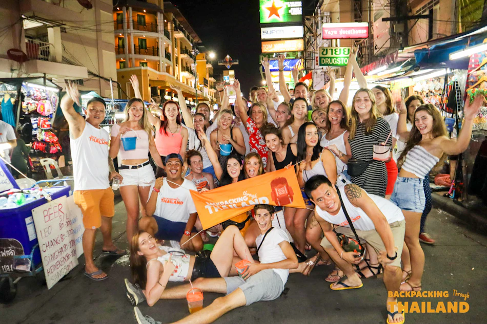 Joyful backpackers celebrate together on a lively, brightly lit street market in Thailand at night.