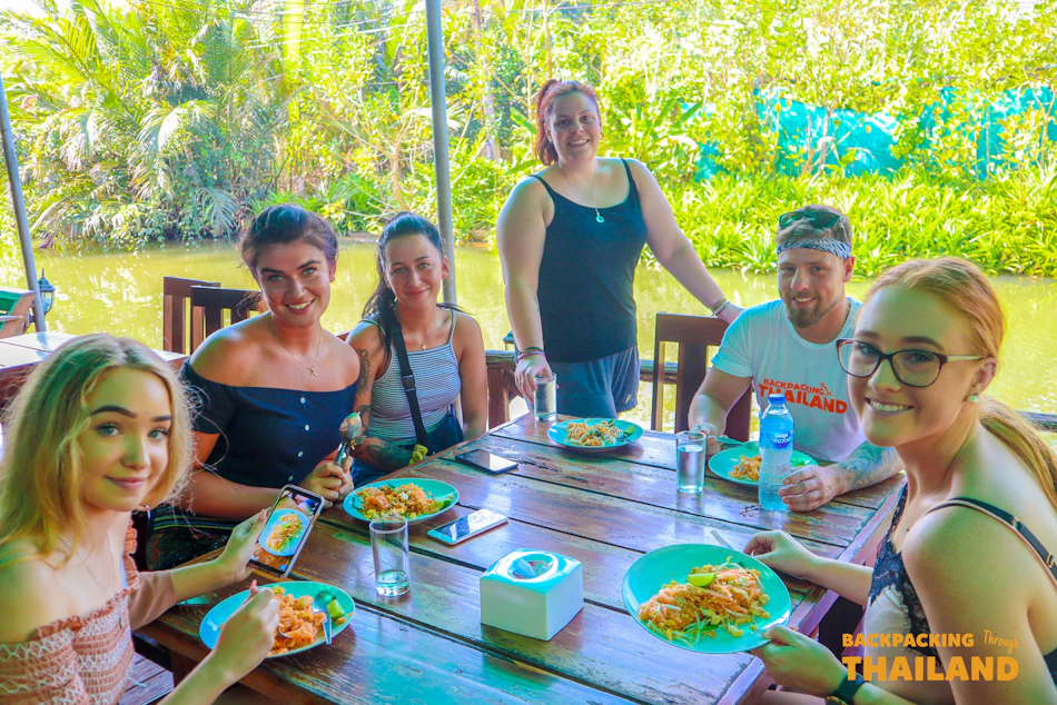 Six happy friends share a delicious meal at an outdoor restaurant by a river in Thailand.