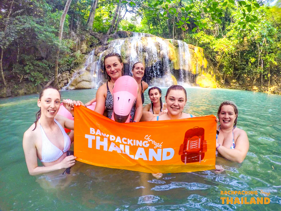 Six smiling women hold a "Backpacking through Thailand" banner in a beautiful jungle waterfall pool.