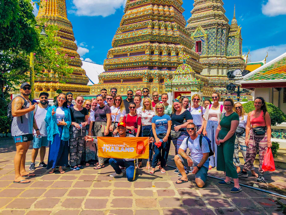 A large group of happy backpackers posing in front of ornate golden temple spires in Bangkok.