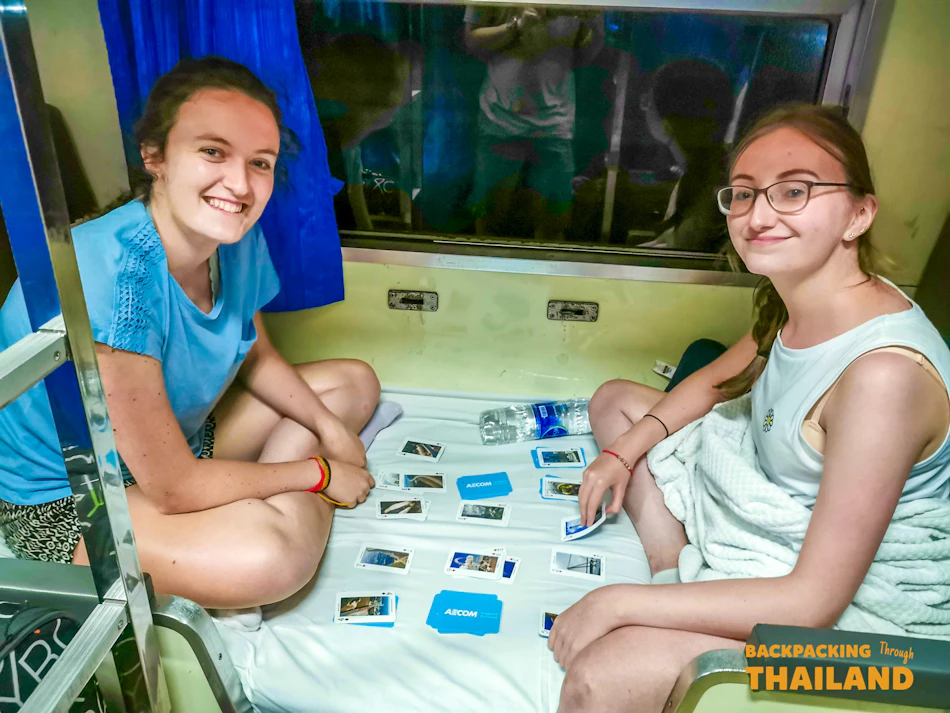 Two smiling women playing cards on a bunk bed inside a night train in Thailand.
