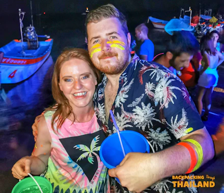 Smiling couple with neon face paint and glow bracelets holding buckets at a Full Moon Party in Thailand.