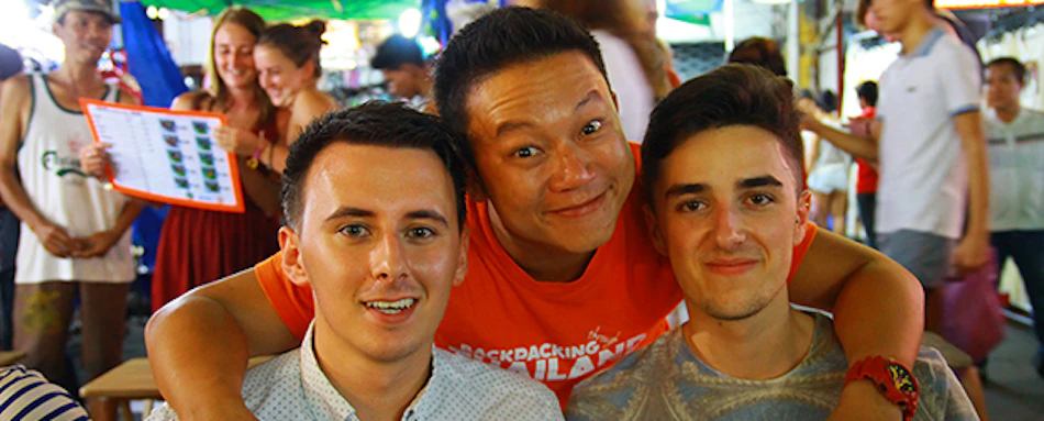 Three friends, one in a "Backpacking Thailand" shirt, smiling happily in a bustling market.