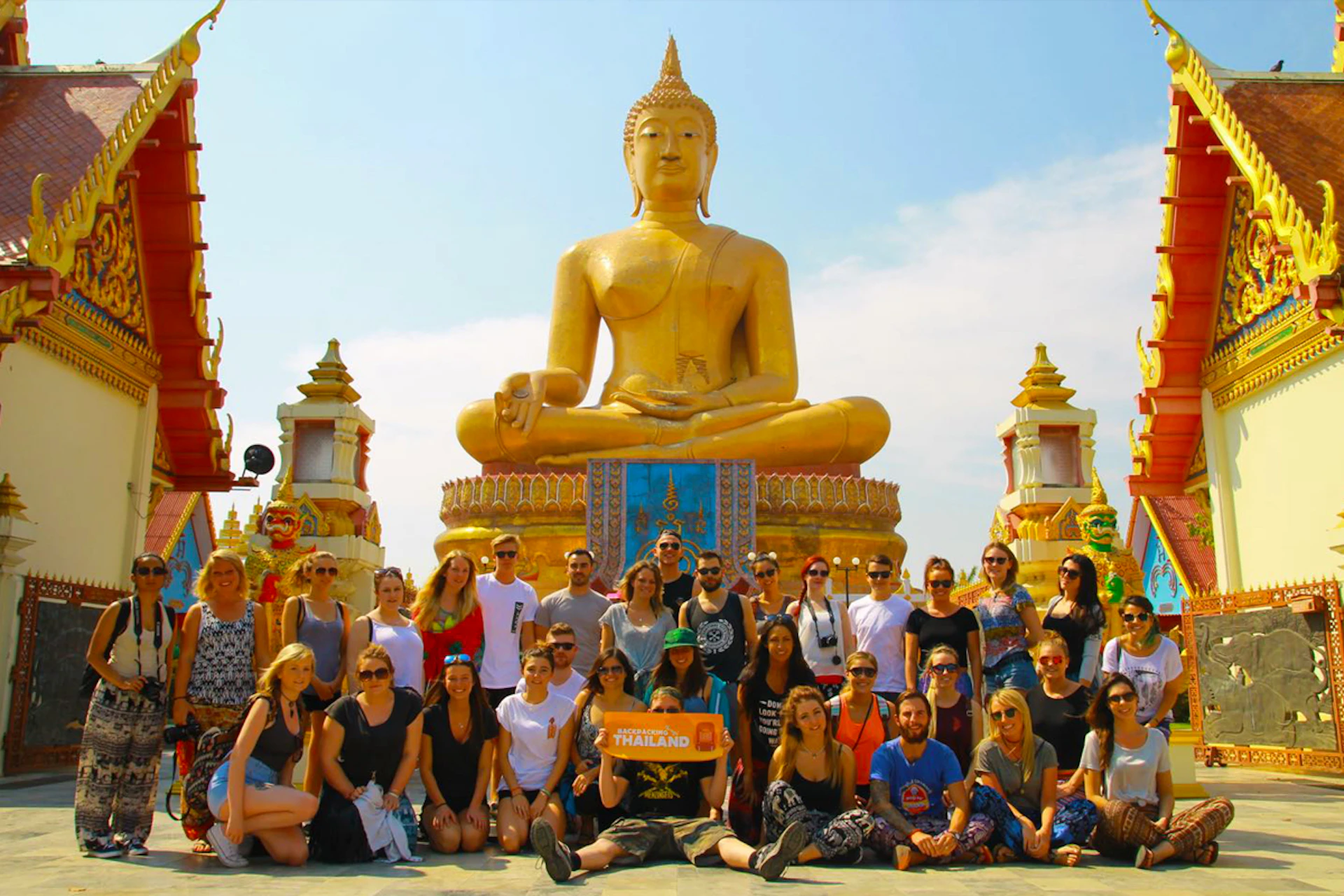 Backpackers posing happily in front of a majestic golden Buddha statue at a temple in Thailand.