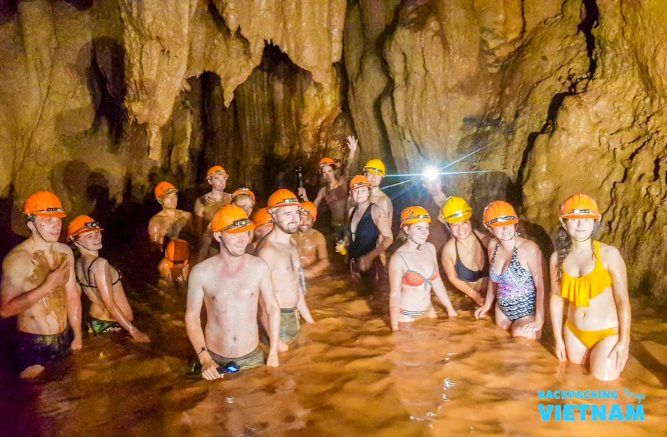 Smiling backpackers in hard hats wading through muddy cave water during an adventure tour in Vietnam.