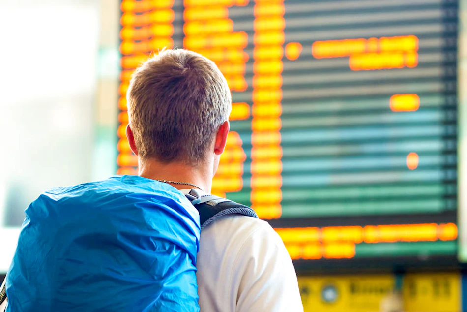 A traveler with a blue backpack stands in an airport, checking flight information on a large board.