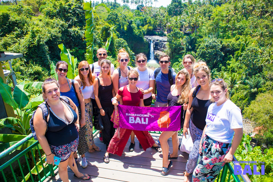 A group of smiling backpackers holding a Bali banner in front of a tropical waterfall.