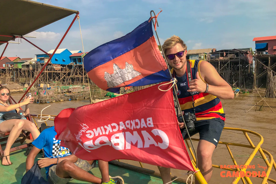 Happy backpacker gives a thumbs-up on a boat in Cambodia with flags and stilt houses.