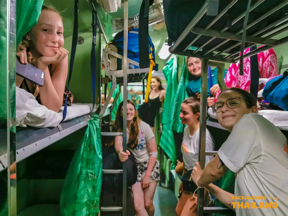 Smiling backpackers relaxing in bunk beds on a lively overnight train journey through Thailand.