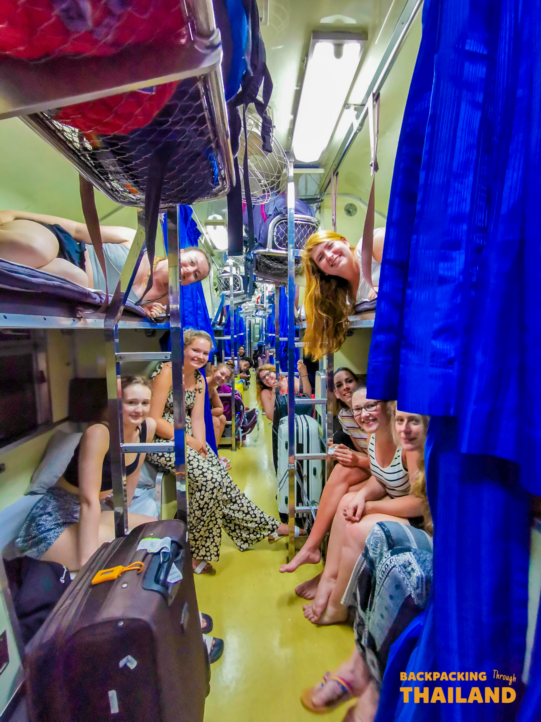 Backpacking travellers sitting in the aisle of an overnight train with luggage and blue curtainsBackpacking travellers smiling and chatting in a train car corridor with green frames and open windows