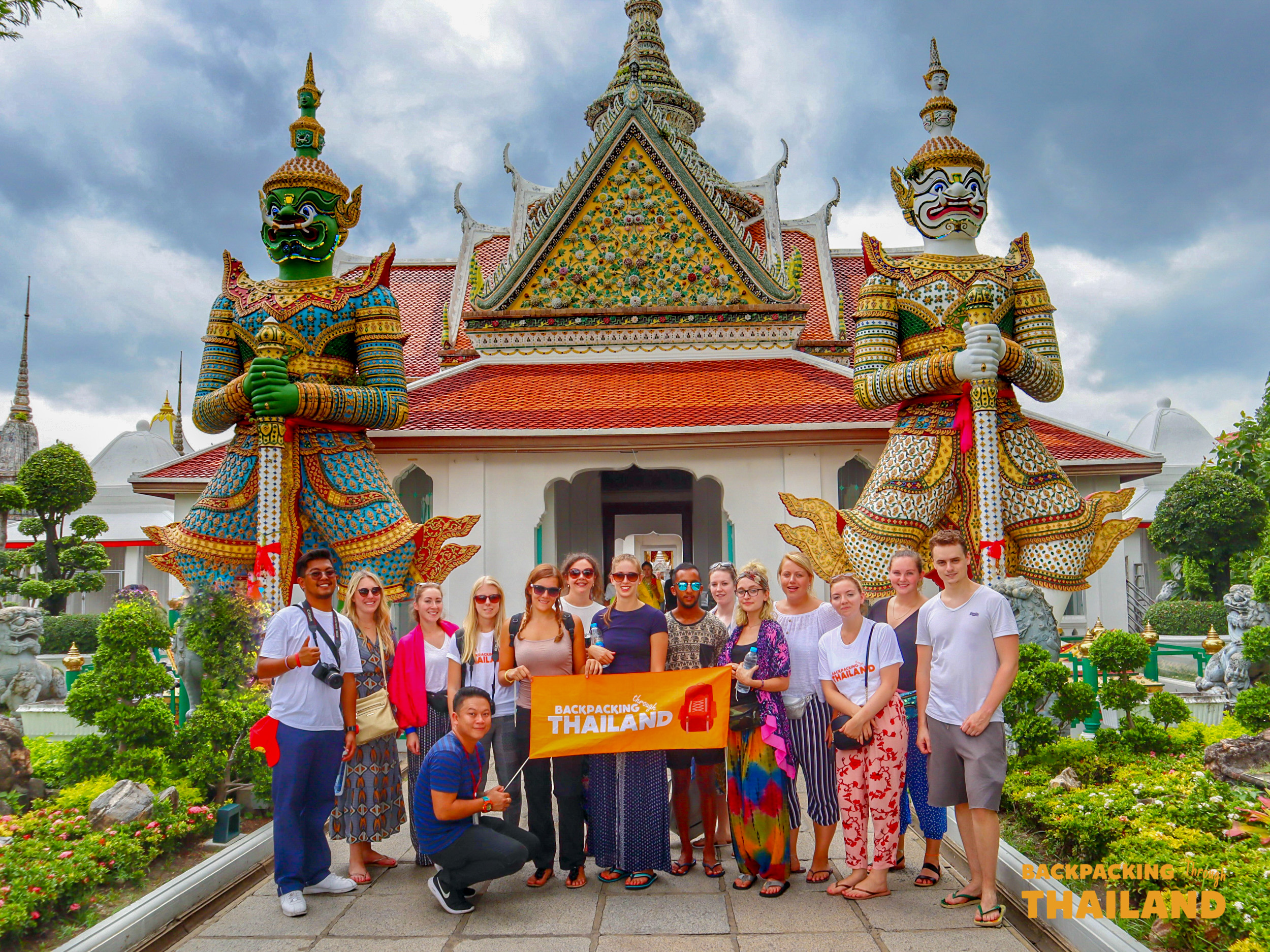 Happy backpackers pose with their tour banner before the giant guardian statues at Wat Arun, Bangkok.