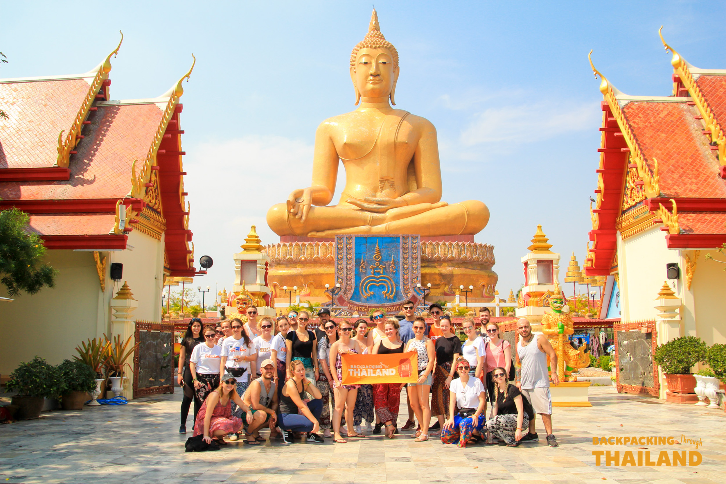 Happy backpackers gather for a group photo before the impressive golden Buddha statue in Singburi, Thailand.