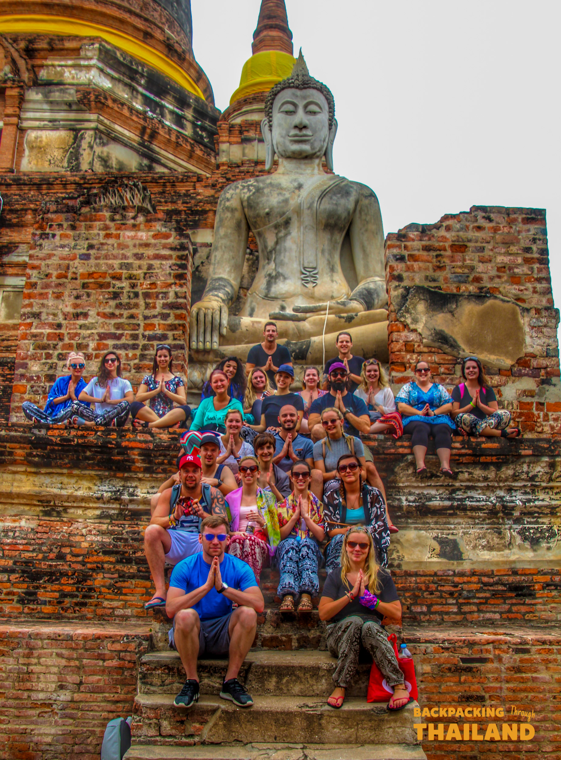 Backpackers pose reverently before a massive Buddha statue amidst the ancient ruins of Ayutthaya.