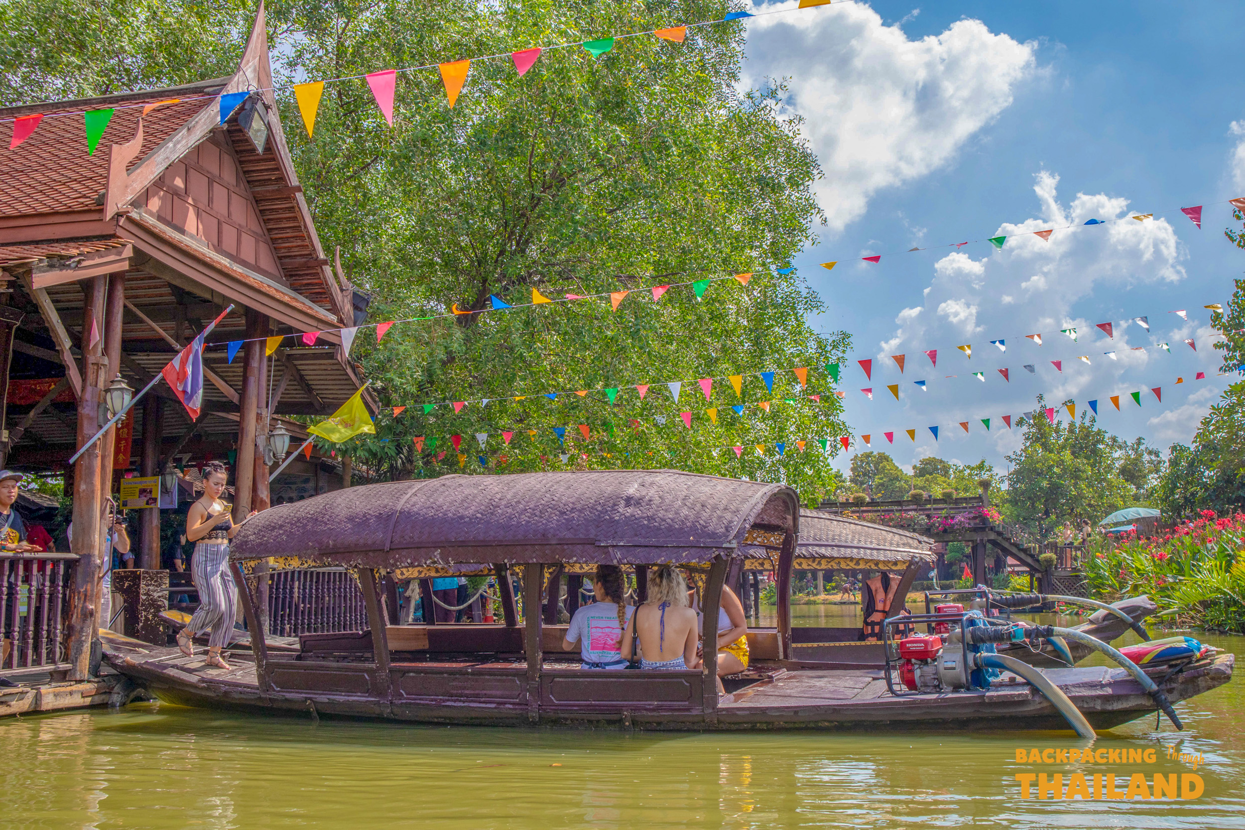 Visitors enjoy a scenic boat ride past traditional Thai buildings and festive flags under a blue sky.