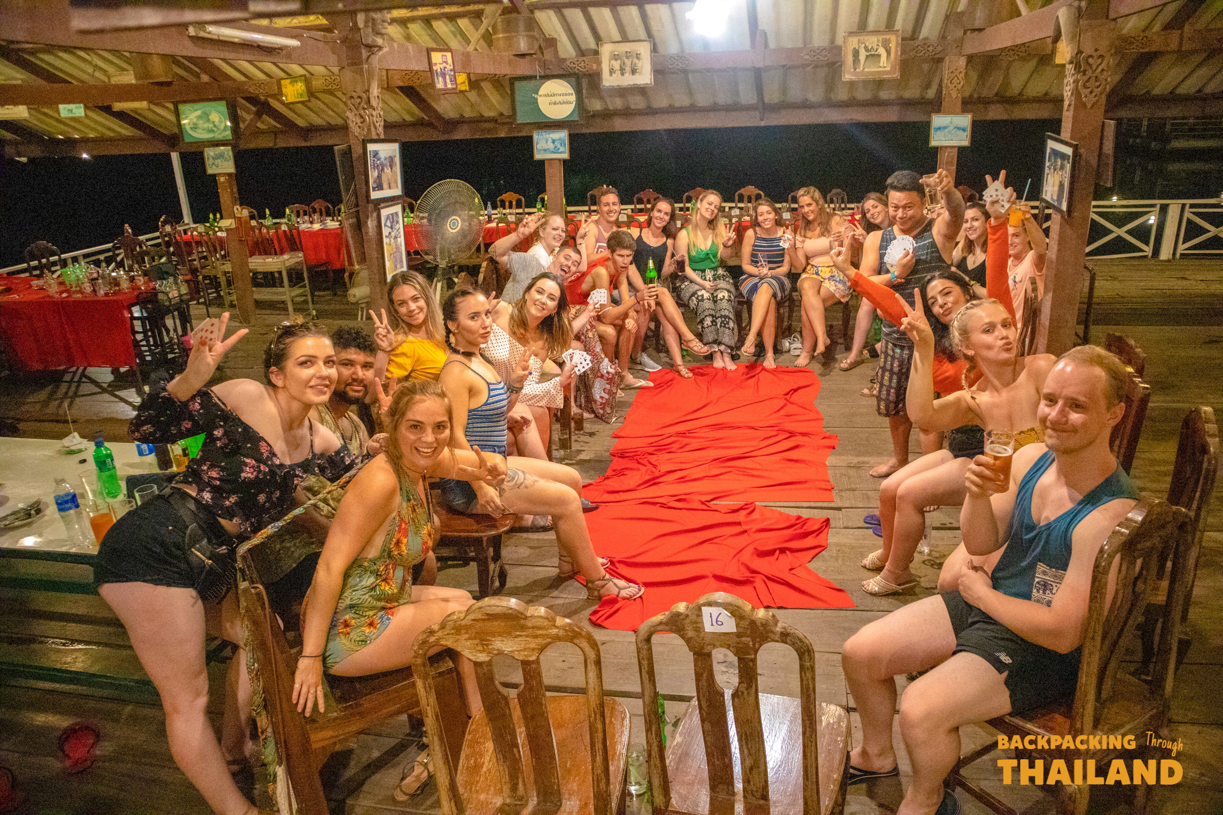 A large group of smiling backpackers enjoys a lively evening in a rustic open-air restaurant in Singburi.