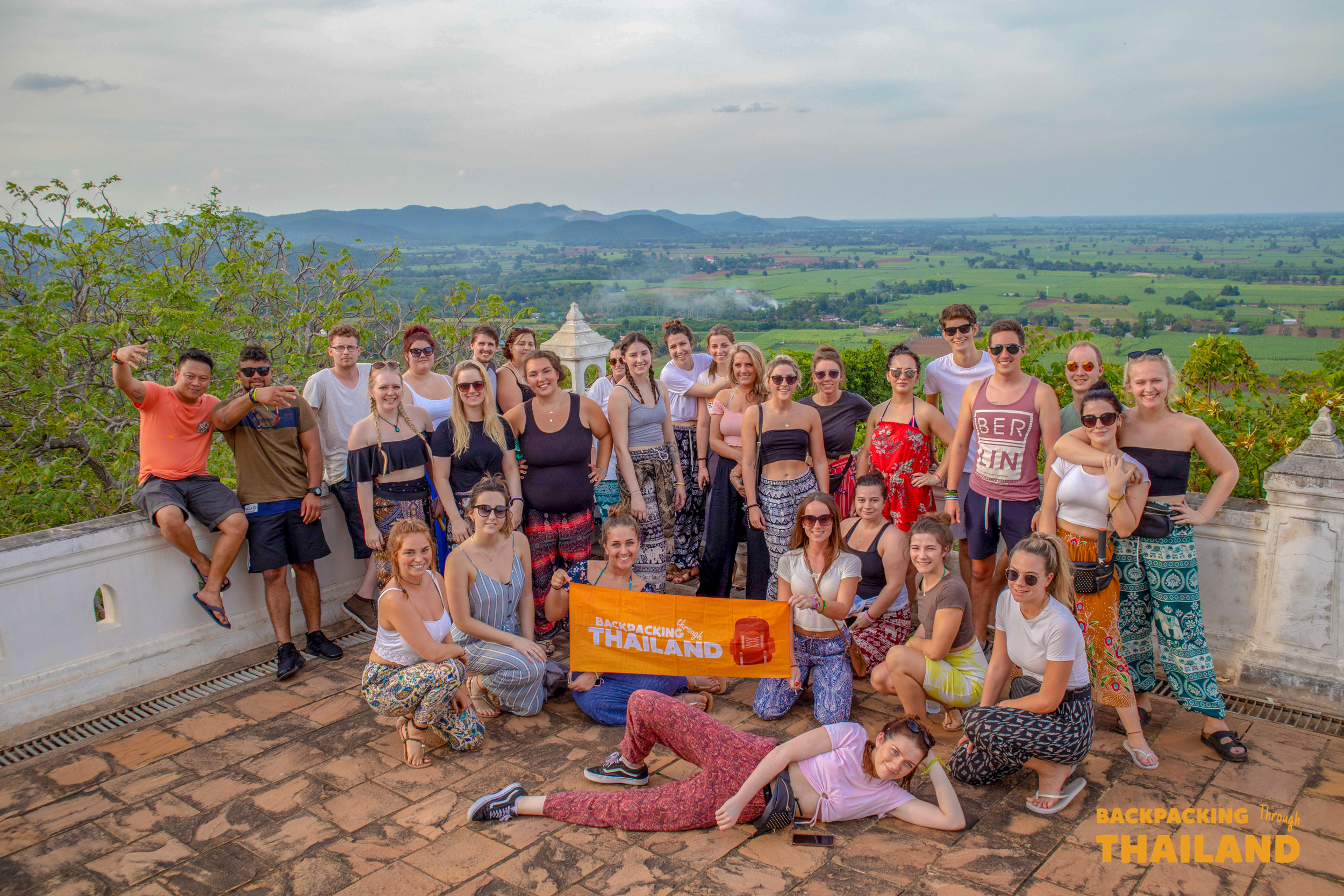 A large group of happy backpackers pose with their "Backpacking Thailand" banner, overlooking scenic Singburi.