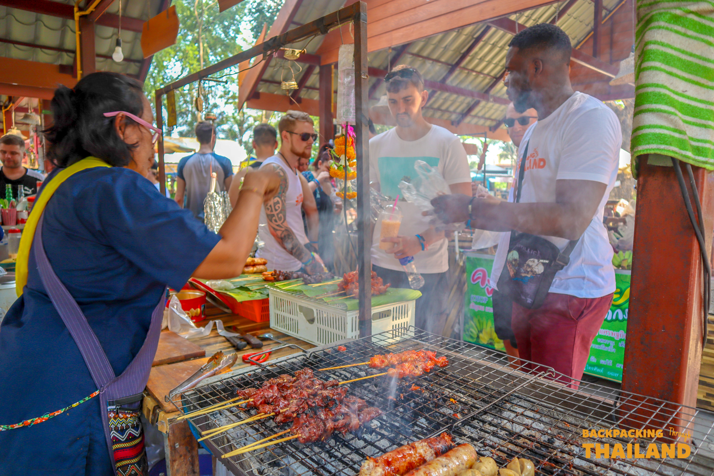A local vendor serves grilled skewers to eager tourists at a vibrant market in Singburi.