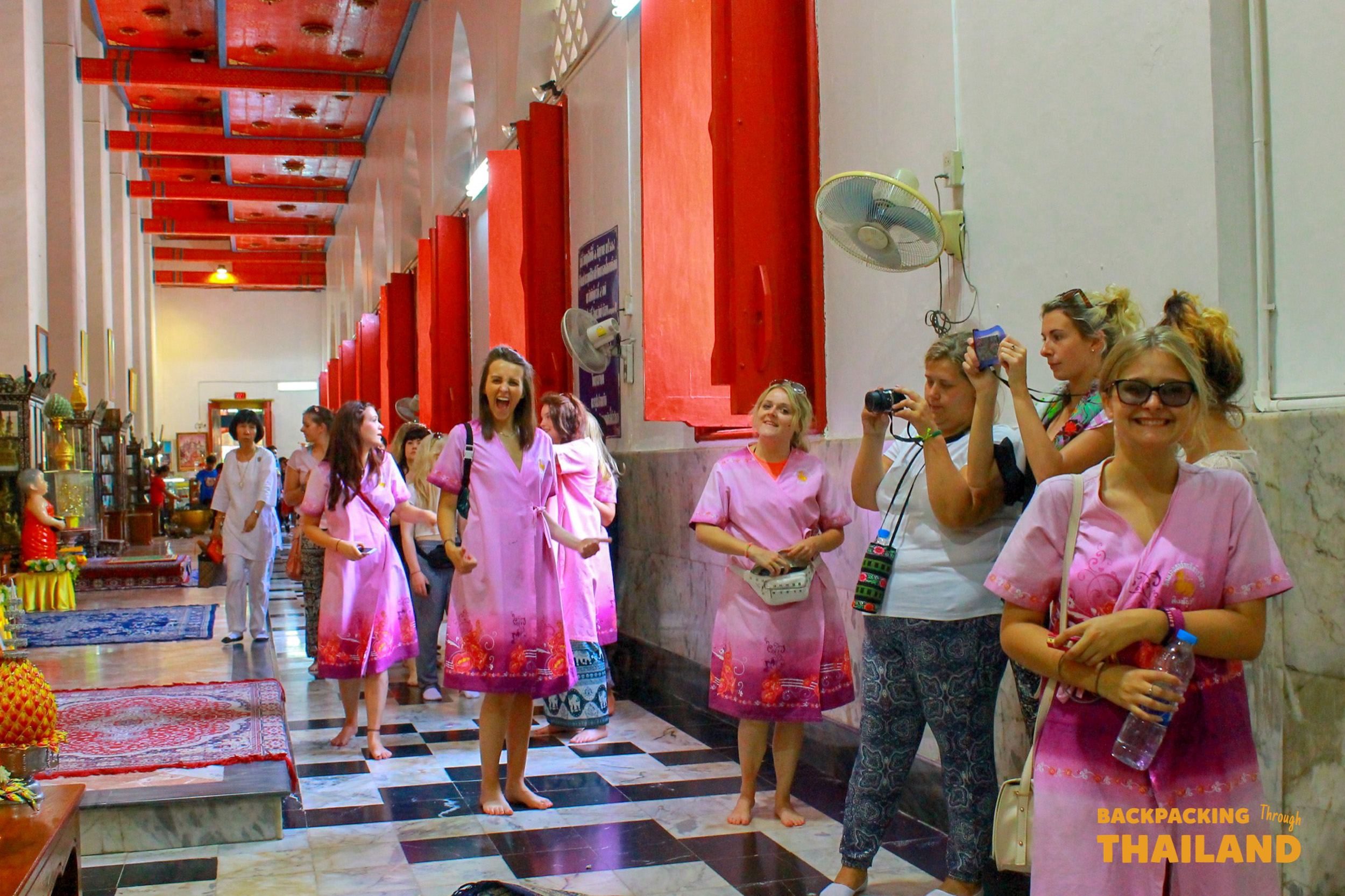 Smiling women in pink dresses explore a vibrant temple hallway, observing the sights in Singburi, Thailand.