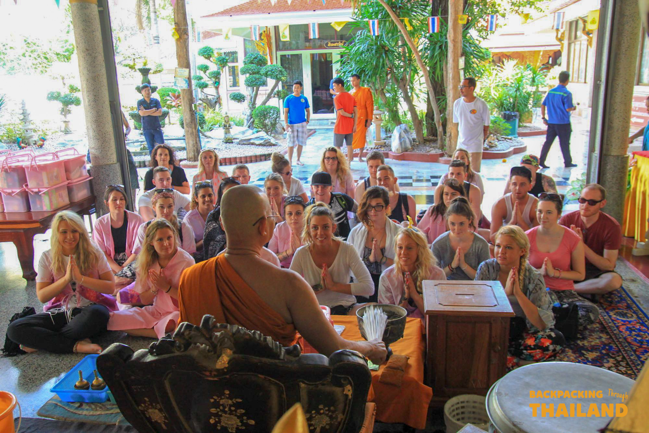 Backpackers respectfully engage with a Buddhist monk during a cultural experience in Singburi, Thailand.