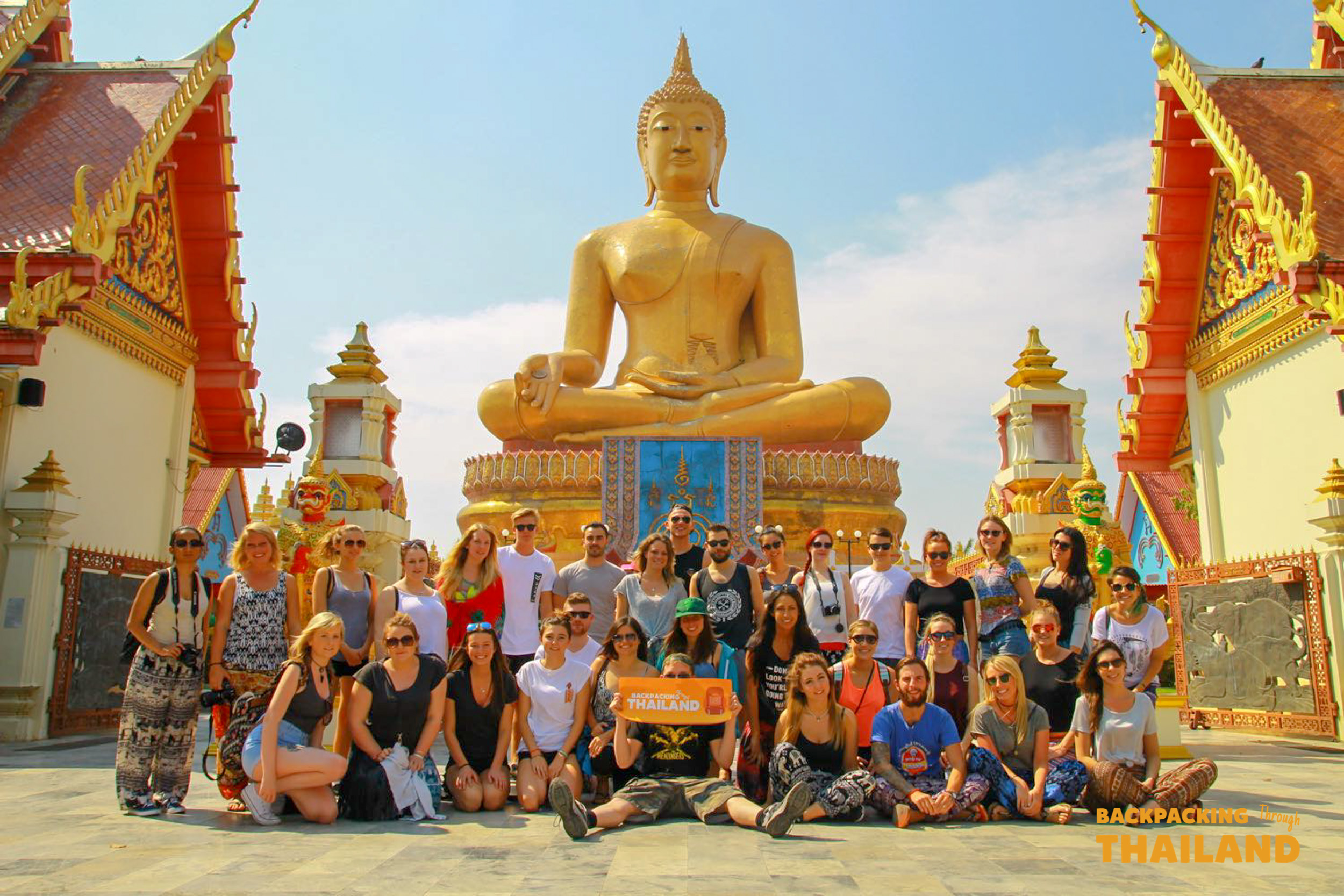 A group of happy backpackers pose in front of the magnificent golden Buddha statue in Singburi, Thailand.