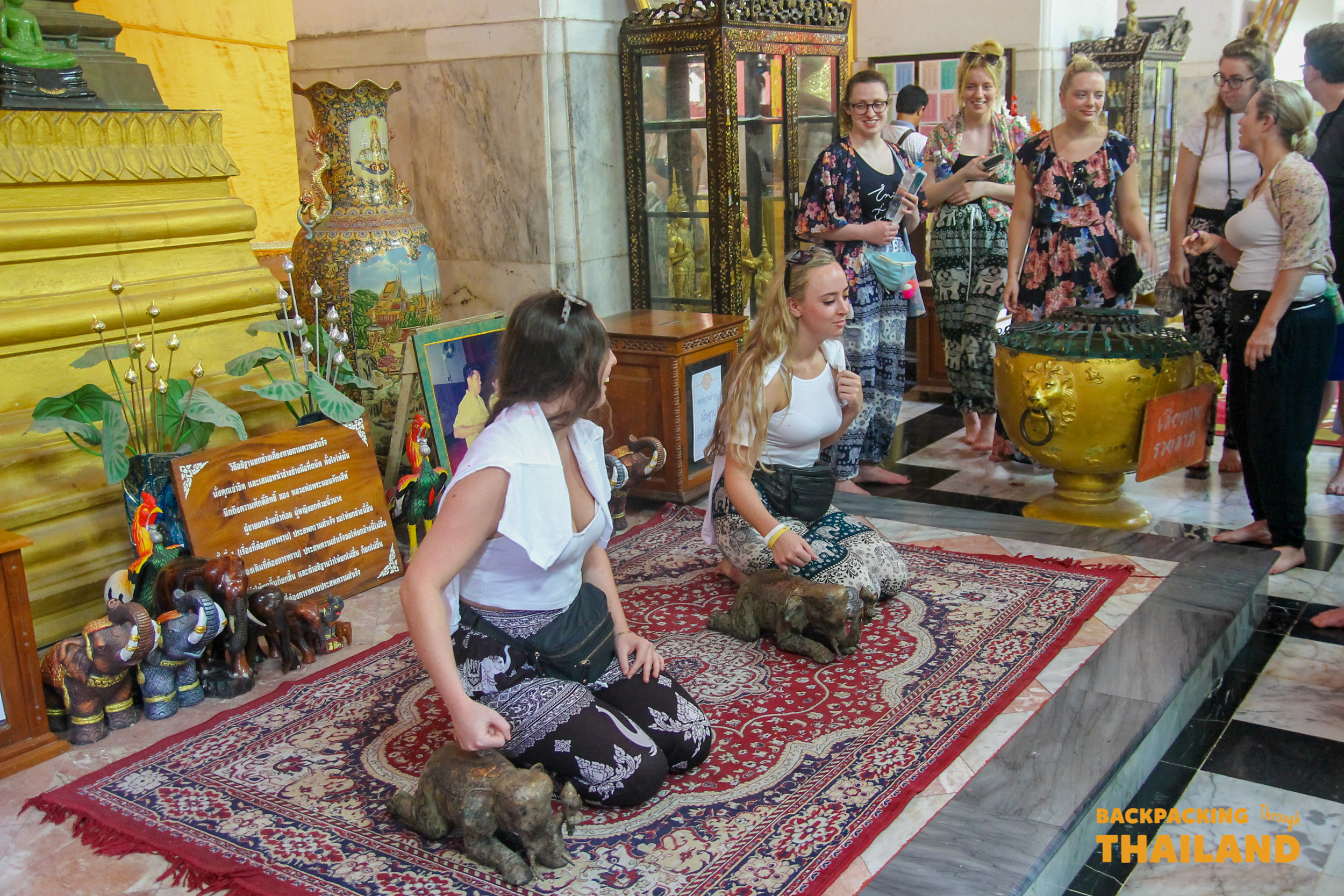 Travelers respectfully kneeling on a rug, touching small statues inside a temple in Singburi.