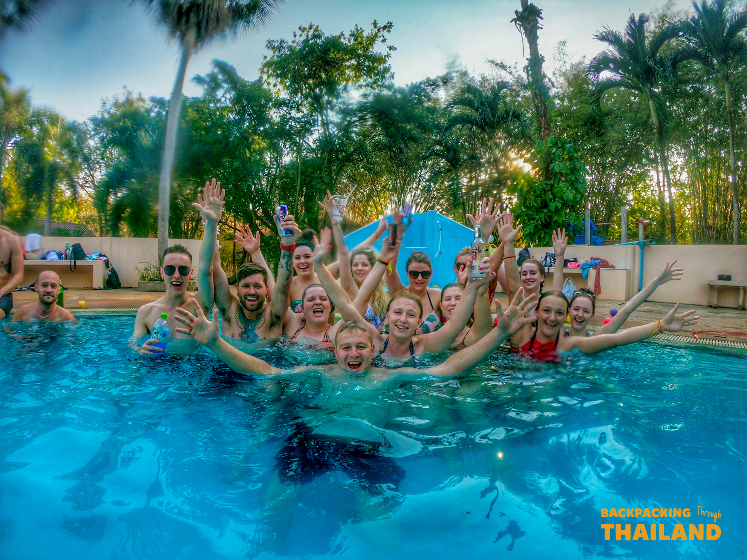 A group of happy backpackers waving from a swimming pool during a lively pool party in Singburi, Thailand.