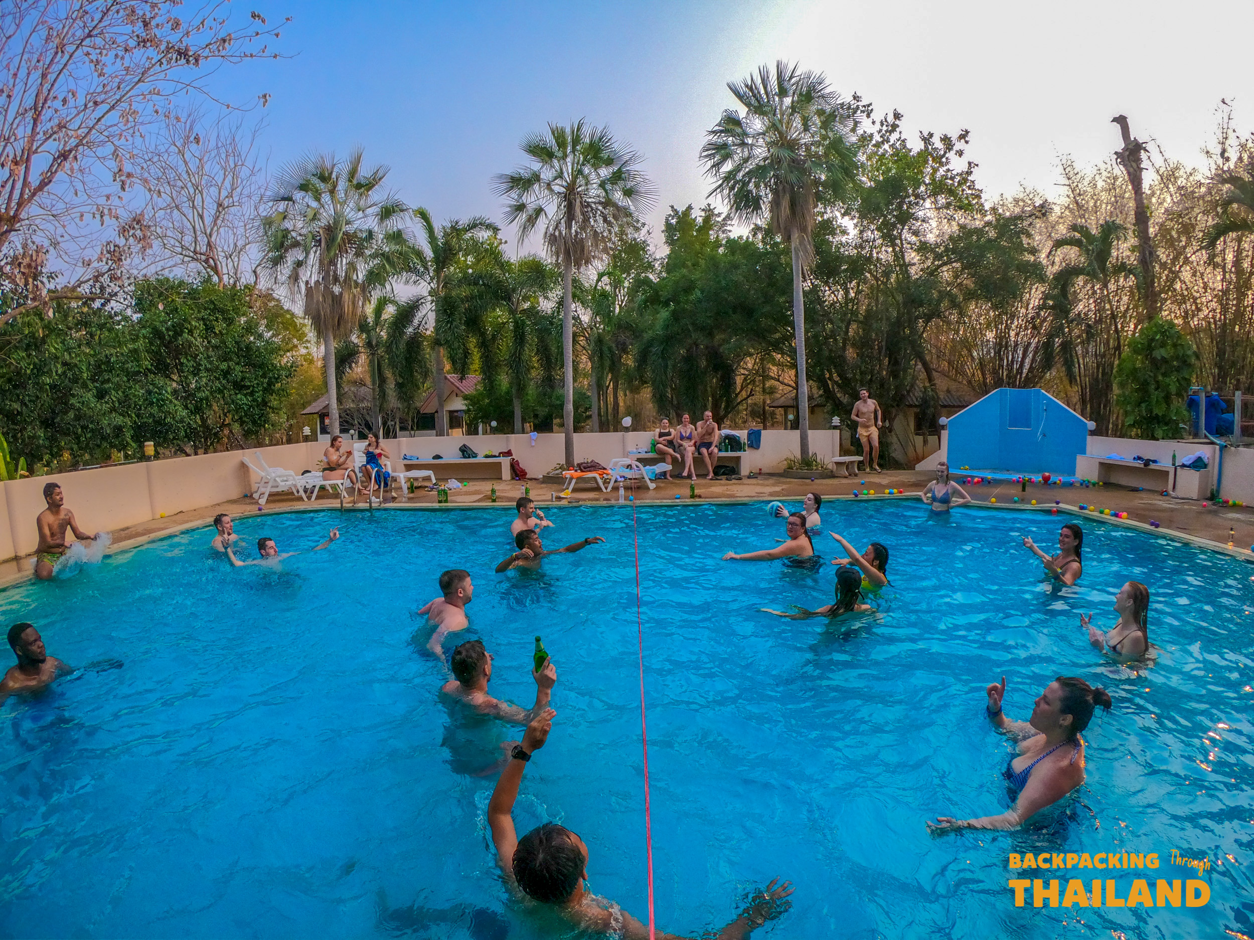 Backpackers enjoy a lively pool party with drinks and games under palm trees in Thailand.