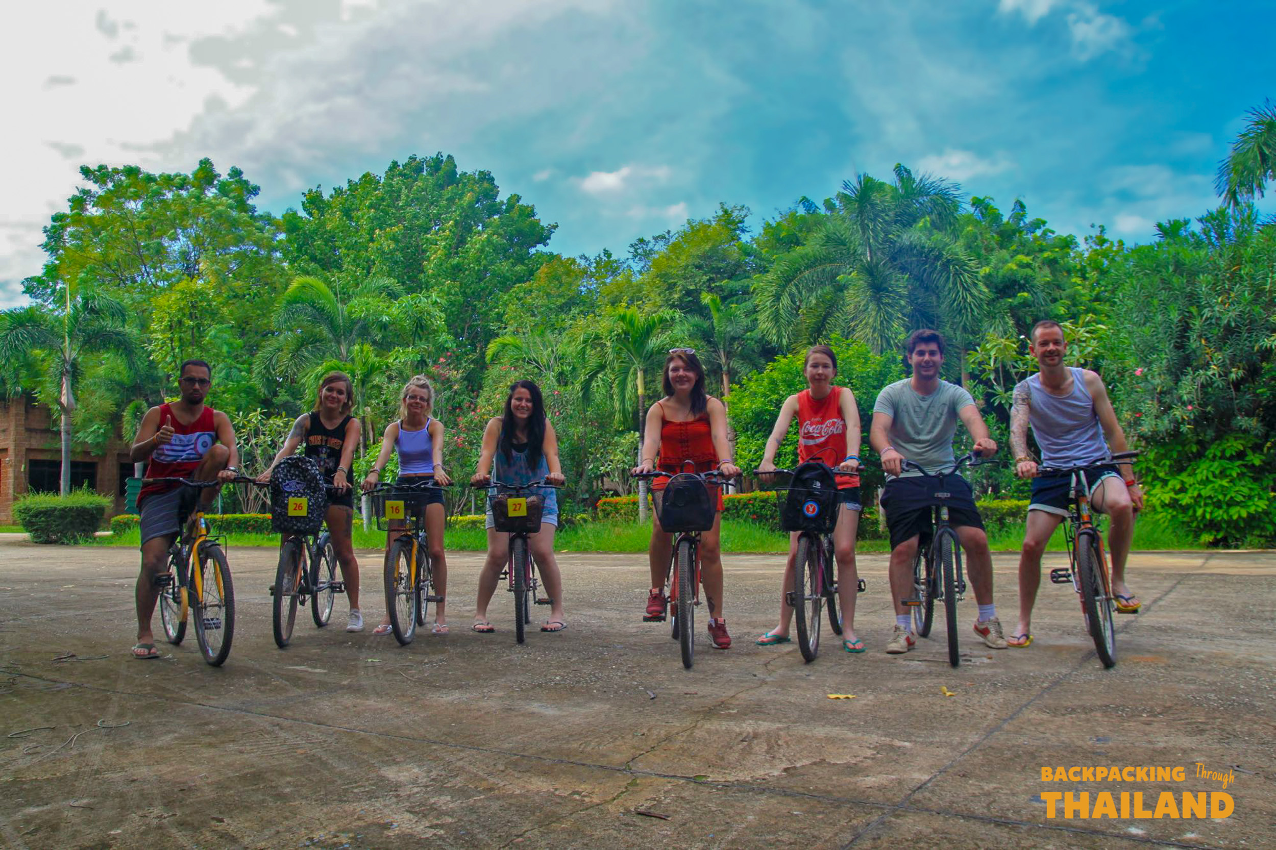 Seven happy backpackers pose with their bicycles on a paved path surrounded by lush greenery in Thailand.