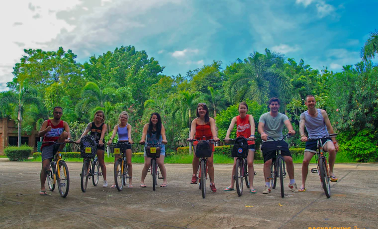 Seven happy backpackers pose with their bicycles on a paved path surrounded by lush greenery in Thailand.